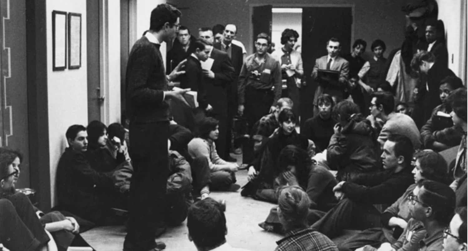 black and white image of bernie sanders at a sit-in in 1962, addressing a group of about 25 people standing and sitting on the floor